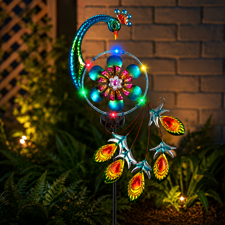 Leuchtender Solar Pfau Gartenstecker mit bunten LEDs als dekorative Gartenbeleuchtung neben Blumenbeet und Terrasse bei Nacht.
