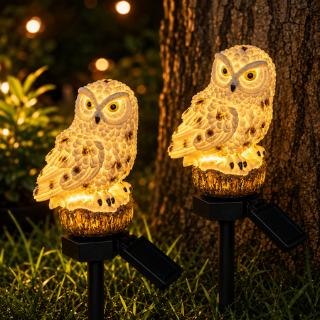 Zwei leuchtende Solar-Eulen als Gartenstecker neben einem Baum mit warmweißem LED-Licht.
